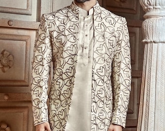 May include: Cream-colored formal suit featuring a jacket with a floral pattern in dark brown. The suit includes a long tunic and matching pants. The jacket has a mandarin collar and is worn over a button-down shirt.