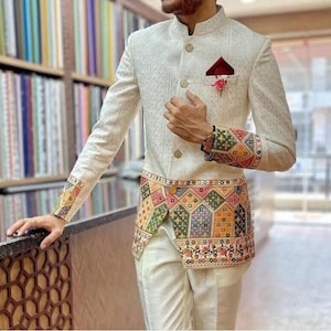 Könnte beinhalten: Elfenbeinfarbener Anzug mit aufwendiger geometrischer Stickerei. Die Jacke hat einen Stehkragen, goldene Knöpfe und ein dekoratives Einstecktuch. Manschetten und Taille sind mit farbenfrohem, gemustertem Stoff verziert. Der Anzug wird mit einer passenden elfenbeinfarbenen Hose kombiniert.