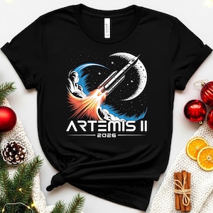 Op de afbeelding: Zwart t-shirt met een afbeelding van een raket die naar de maan lanceert, met de tekst "ARTEMIS II 2026". Het ontwerp bevat oranje en blauwe vlammen, een sterrenhemel en een halve maan.