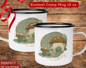 May include: Two white enamel camp mugs with black rims. The mugs feature a whimsical illustration of a mushroom house with a frog sitting on a toadstool. One mug says "ashley" on the bottom. The mugs are 12 ounces.