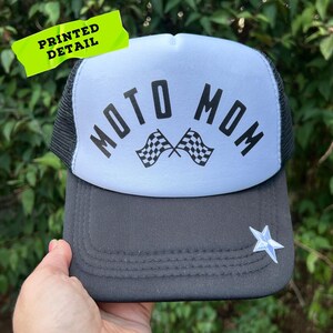 May include: A trucker hat with a white front panel and a black mesh back. The hat features the text "MOTO MOM" above two checkered flags. A star-shaped patch is on the brim. The words "PRINTED DETAIL" are on a neon green sticker.