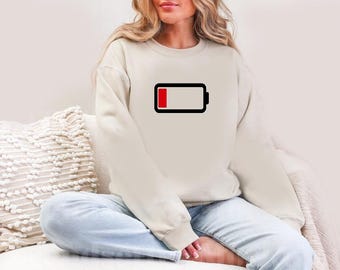 Puede incluir: Una sudadera de color crema con un gráfico de icono de batería baja en negro y rojo.