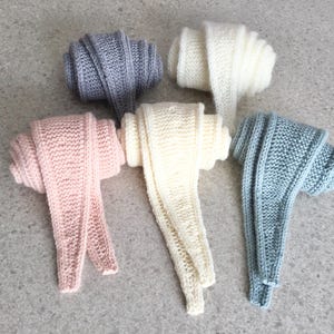 May include: Five rolled knit scarves in various colors: light pink, off-white, light blue, gray, and white. The scarves are made of a chunky knit material and are displayed on a gray surface. The scarves are neatly rolled.