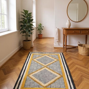 May include: A rectangular rug with a geometric diamond pattern. The rug features a black border, a yellow diamond design, and a striped gray and white background. The rug has a fringe along the edges and is placed on a wooden floor.
