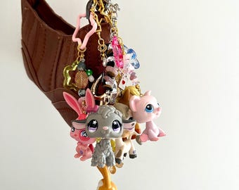Puede incluir: Un bolso de hombro marrón adornado con un llavero de varias capas que presenta diversas figuras de animales de plástico, incluyendo un cordero gris, un gato rosa y un pequeño perro naranja y blanco, junto a dijes de estrellas, cuentas y metal.