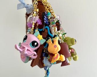 Puede incluir: Un colgante para bolso marrón que presenta una colección de figuras de animales de plástico de colores, incluyendo un pulpo rosa, un caracol naranja y un pez azul. El colgante está decorado con cadenas doradas, cuentas, estrellas y una cinta azul.