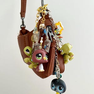 Peut inclure: Un sac à bandoulière en similicuir marron orné d'une collection de breloques suspendues. Les breloques comprennent des figurines d'animaux en plastique, des perles en acrylique transparent, des chaînes dorées et des étoiles fixées à la fermeture.