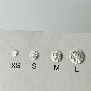 May include: Four silver stud earrings in varying sizes, labeled XS, S, M, and L. The earrings have a hammered texture and are displayed on a white surface. The sizes range from small to large, offering a selection for different preferences.