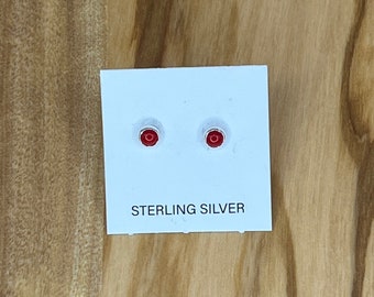 May include: A pair of small, round, red stud earrings on a white card. The card is labeled "STERLING SILVER" in black text. The earrings are set against a light wood grain background.