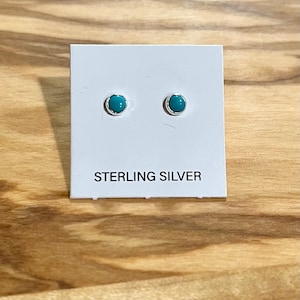 May include: A pair of sterling silver stud earrings with round, turquoise-colored stones. The earrings are set in a simple, polished silver bezel. The card beneath the earrings reads "STERLING SILVER".