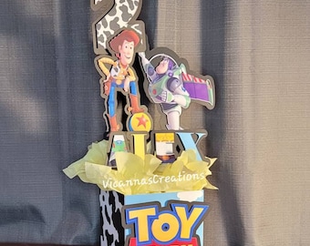 May include: A Toy Story-themed centerpiece featuring Woody and Buzz Lightyear figures, the number 2, and the name "ALEX". The box has a cow print design and the words "TOY STORY". Yellow tissue paper fills the box.