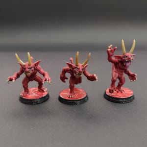 Op de afbeelding: Drie miniatuur rode demonen beeldjes met gele hoorns en scherpe tanden. Elke figuur poseert anders, staand op een rode cirkelvormige basis met een zwarte rand. De figuren hebben een dreigende uitstraling, met uitgestrekte klauwen en een grimas.