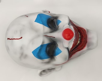 May include: A white creepy clown mask with blue and red paint. The mask has a wide, toothy grin and a red nose.