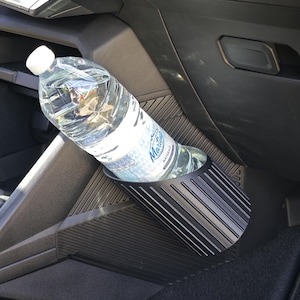 Pode incluir: Um suporte para copos preto, cilíndrico e canelado, montado no painel interior de um automóvel, contendo uma garrafa de água de plástico transparente com tampa branca e um rótulo com o texto Marti e Naturale em azul e branco.