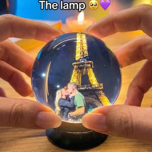 May include: A clear glass snow globe featuring the Eiffel Tower and a couple kissing. The base is black. The text "The lamp" is at the top. A lit candle is in the background.