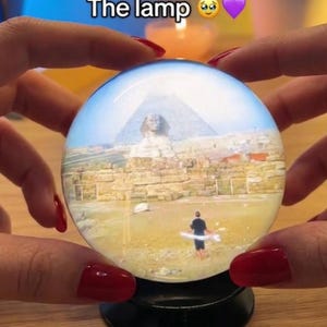 May include: A glass sphere featuring an image of the Sphinx and a pyramid. The sphere is held by hands with red nail polish. The text "The lamp" is visible at the top of the image.