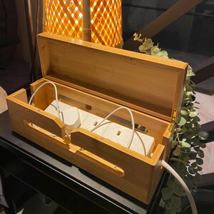 May include: A wooden box with a hinged lid, designed to organize and conceal cords and power adapters. The box has a light wood finish and features three electrical outlets.