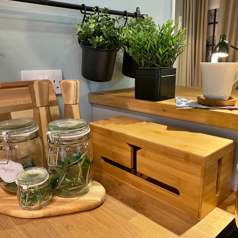 May include: A wooden cable management box with a natural finish. The box has a slot for cords and is sitting on a wooden countertop. There are three glass jars with green herbs and a wooden cutting board in the background.