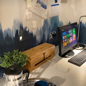 Puede incluir: Una caja de gestión de cables de madera con un cable blanco que sale de ella. La caja está situada en una encimera blanca junto a un monitor de ordenador y un teclado. Hay una planta en maceta y una jarra de vidrio en la encimera.