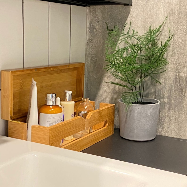 May include: A bamboo bathroom organizer with a lid, holding various bottles of toiletries. The organizer is sitting on a black countertop next to a potted plant with green leaves.