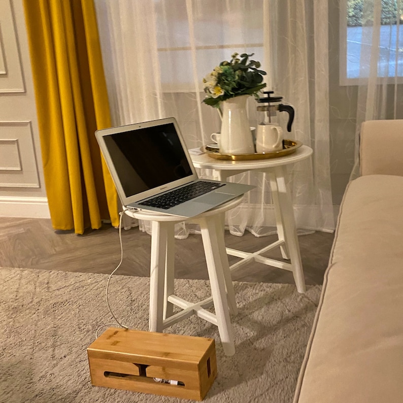 May include: A white laptop computer sits on a white table with a wooden cable organizer. The table is next to a white couch and a yellow curtain.