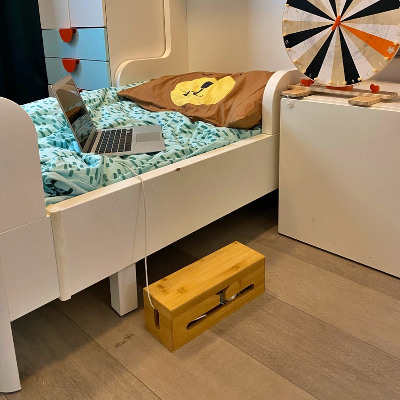 May include: A white wooden bed frame with a blue and green patterned bedspread. A laptop computer is on the bedspread. A wooden cord organizer is on the floor under the bed.