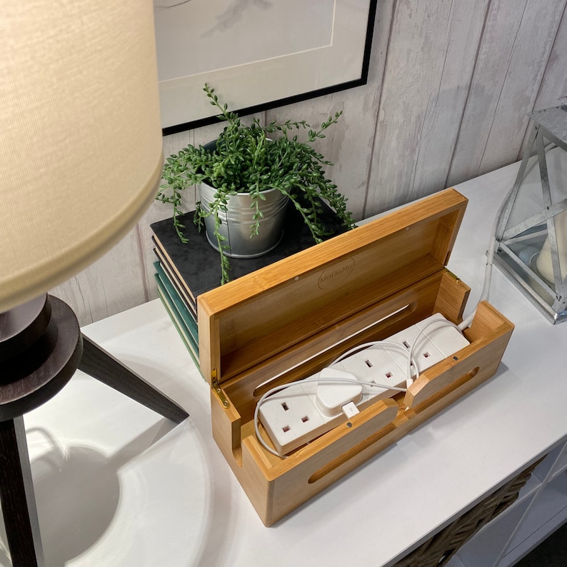 May include: A bamboo cord management box with a lid open, revealing a white power strip with multiple outlets. The box is sitting on a white surface with a plant in a green pot and a stack of books.