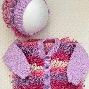 May include: A knitted baby cardigan and hat set in shades of pink, purple, and white. The cardigan has a button-down front and long sleeves. The hat has a textured trim. The set is handmade, with a fluffy, textured yarn.