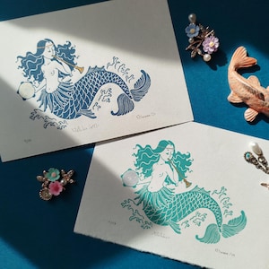Peut inclure: Deux impressions d'une sirène jouant de la flûte. La sirène sur l'impression du haut a une queue bleue et la sirène sur l'impression du bas a une queue turquoise. Les deux impressions ont un fond blanc.