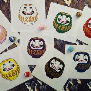 May include: Eight colorful Daruma dolls, a traditional Japanese good luck charm, with gold accents and the kanji for "luck" on each. The dolls are on white paper with small decorative charms.