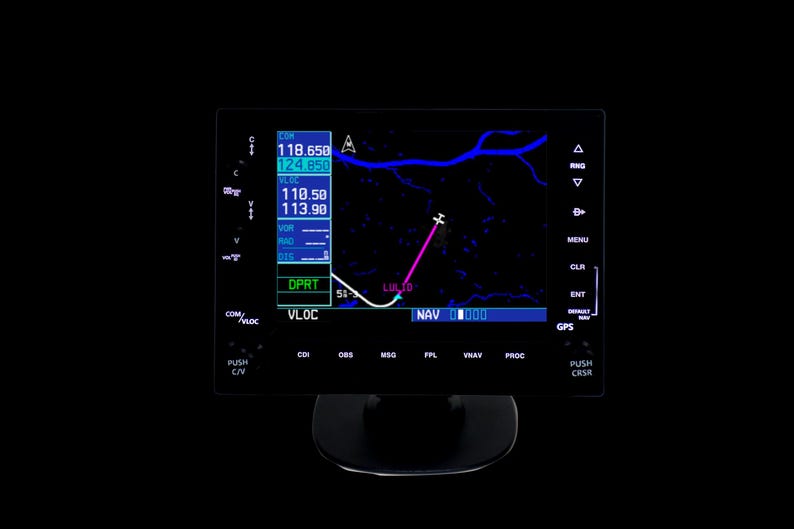 May include: A black aviation instrument panel displaying flight data, including frequencies and navigation information. The screen shows a flight path with a pink line and the text "LUL TO". The panel has various buttons and labels.