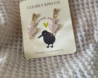 May include: A pair of pink heart-shaped dangle earrings with gold accents. The earrings are handmade and feature a small kiwi bird charm. The card reads "CLEARLY.KIWI.CO HANDMADE JEWELRY ETSY SHOP: CLEARLYKIWICLAY"