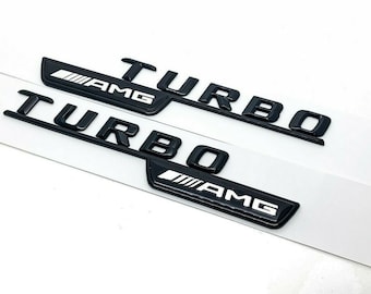 May include: Two black "TURBO" and "AMG" emblems. The emblems are glossy black with white lettering. The AMG emblem has a white stripe design. The emblems are on a white background.