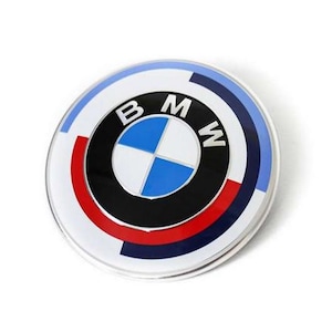 May include: Circular emblem featuring the BMW logo. The design includes a white outer ring, a blue and white checkered center, and the letters "B", "M", and "W" in black. Red and blue accents are also present.