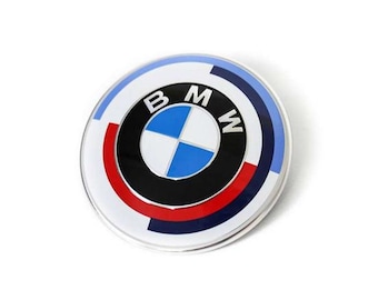 May include: Circular emblem featuring the BMW logo. The design includes a white outer ring, a blue and white checkered center, and the letters "B", "M", and "W" in black. Red and blue accents are also present.