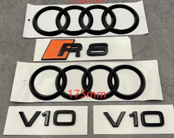 May include: Black car emblems, including the four interlocking rings logo (192mm and 175mm), the R8 logo with an orange background, and two V10 emblems. The emblems are on white backing.