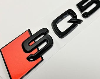 May include: Black "SQ5" emblem with a red and black background. The emblem is glossy and appears to be made of plastic. The letters are thick and bold, with a modern font.
