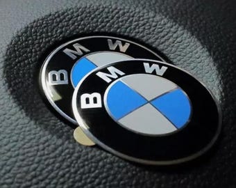 May include: Two BMW emblems, one slightly overlapping the other, are displayed against a textured black surface. The emblems feature a black outer ring with the letters "BMW" in white. The inner circle is divided into blue and white quadrants.