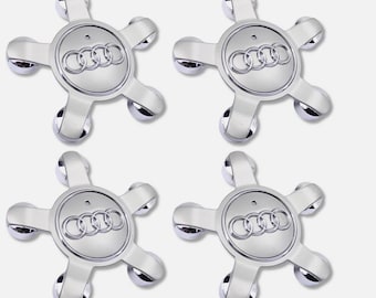 May include: Four silver-colored wheel center caps with the Audi logo. Each cap has a star-like design with five rounded extensions and a circular center. The Audi logo consists of four interlocking rings.