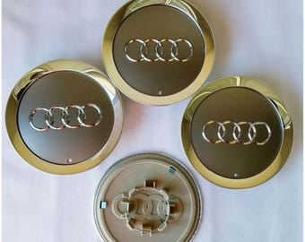 May include: Four silver and gold Audi wheel center caps. The caps have a dark gray center with the Audi logo in silver. The outer ring is gold. One cap is turned over, showing the back.