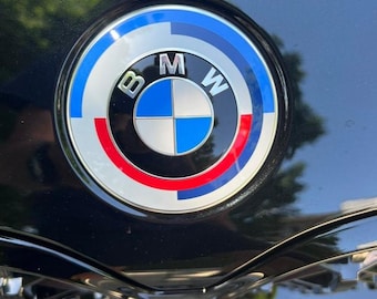 May include: Close-up of a BMW emblem. The emblem is a circular design with a silver border, featuring the letters "BMW" and a blue and white checkered center. The outer ring has blue, red, and white segments.