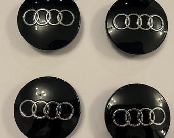 May include: Four black wheel center caps with a silver Audi logo. Each cap is round and glossy, with the Audi rings prominently displayed in the center. The caps are arranged in a square formation.