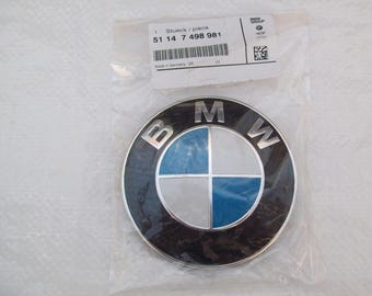 May include: A BMW emblem in a clear plastic bag. The emblem is round with a black outer ring and a silver inner ring. The center features a blue and white cross design, with the letters "BMW" in silver at the top. A white label with text is visible.