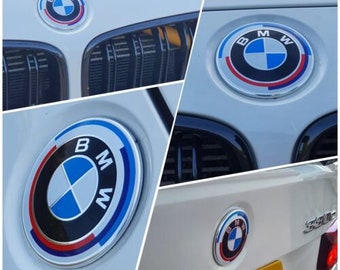 May include: Close-up shots of BMW car emblems. The emblems feature a circular design with the BMW logo in the center, surrounded by blue, black, and red accents. The images show the emblems on the front and rear of a white car.