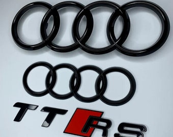 May include: Black car emblem set featuring the Audi rings logo and TT RS lettering. The Audi rings are a set of four interlocking rings. The TT RS lettering is black with a red background.