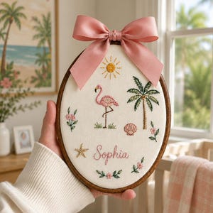 Könnte beinhalten: Ein ovaler Stickrahmen mit weißem Stoffhintergrund und gestickten Motiven eines rosa Flamingos, einer Palme, einer gelben Sonne, einer Muschel, eines Seesterns und kleiner Blumen. Der Name Sophia ist in rosa Schreibschrift gestickt und mit einer rosa Satinschleife verziert.