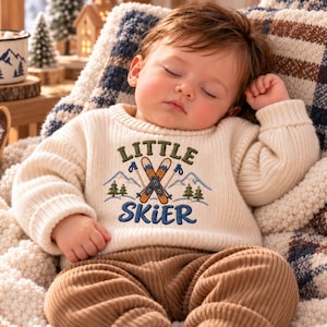 Könnte beinhalten: Ein cremefarbener Langarm-Strickpullover mit einer Stickerei aus zwei gekreuzten Skiern, Bergen und Tannenbäumen sowie dem Schriftzug "LITTLE SKIER" in grünem und blauem Garn, kombiniert mit einer braunen Rippenhose.