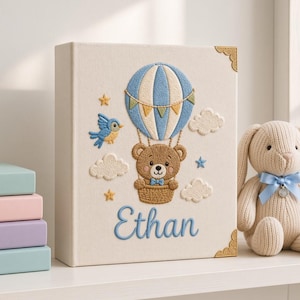 Könnte beinhalten: Ein cremefarbenes Fotoalbum mit Stickereien eines braunen Teddybären in einem blau-weißen Heißluftballon, einem blauen Vogel, Wolken und Sternen. Der Name Ethan ist in blauer Schreibschrift unter dem Ballon gestickt. Goldene Metallecken.