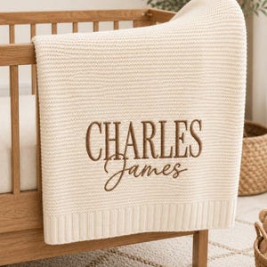 May include: A cream-coloured knitted baby blanket draped over a wooden cot. The blanket features a textured, ribbed knit pattern and is personalised with the name "CHARLES James" embroidered in brown thread in a combination of serif and script fonts.