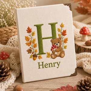 Könnte beinhalten: Ein rechteckiges, cremefarbenes, stoffbezogenes Schild mit einem gestickten grünen Buchstaben H, einem braunen Teddybären, der einen rot-weißen Pilz hält, und Herbstblättern. Der Name Henry ist darunter in Grün gestickt. Dekorative Metallecken.